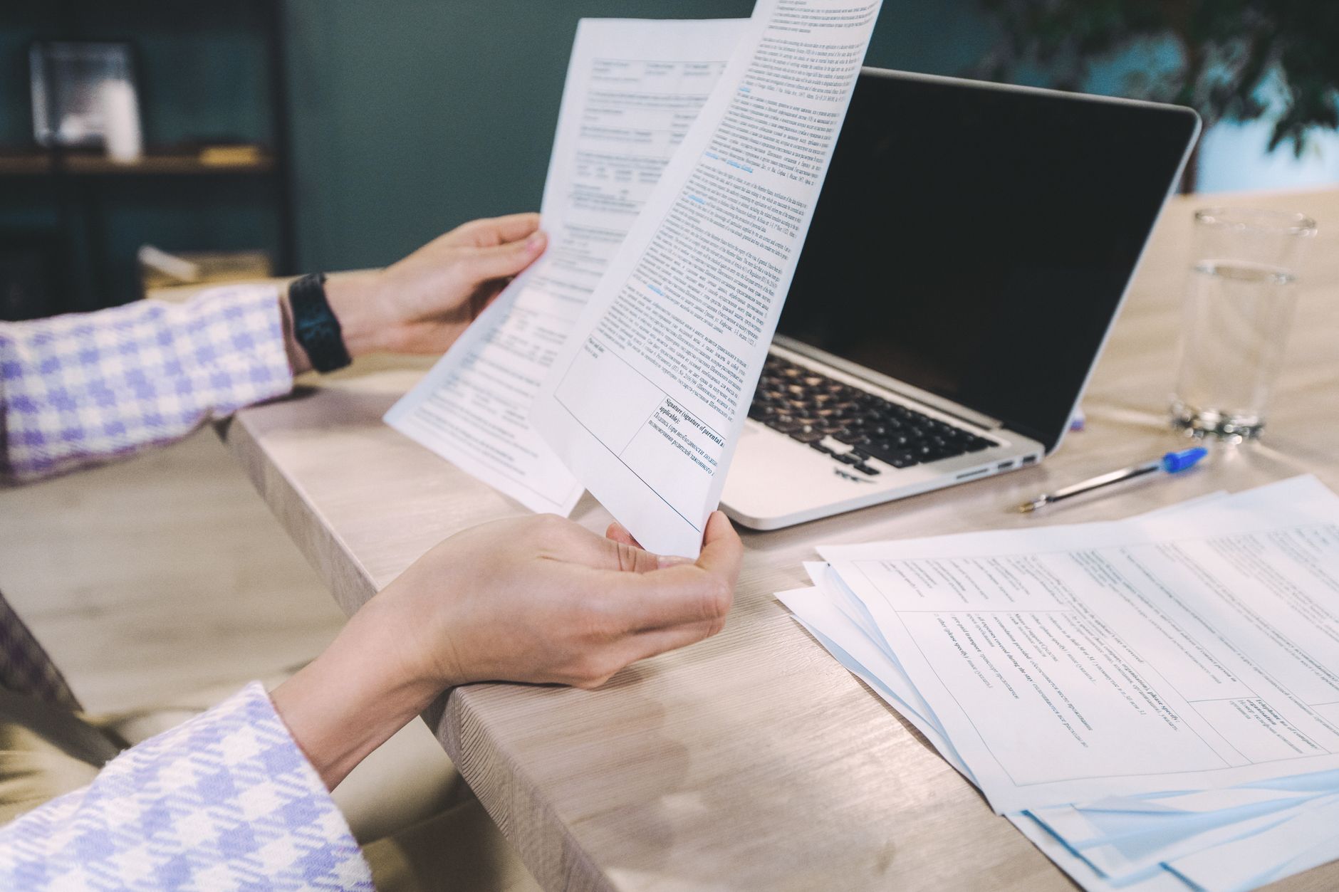 Hands holding a printed policy document at a desk, a second set of papers and a laptop alongside.